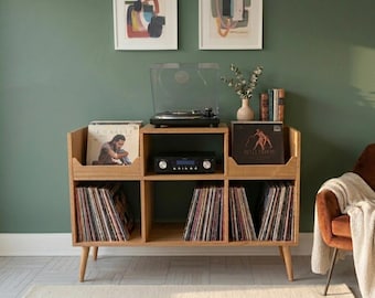 May include: A wooden record player stand with a turntable, amplifier, and vinyl record storage. The stand has a mid-century modern design with angled legs. Two framed abstract art pieces hang on the green wall behind the stand.