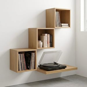 Op de afbeelding: Een moderne wandgemonteerde boekenplank met vier houten kubusplanken. De planken tonen boeken, vinylplaten en een zwarte platenspeler. De unit is gemonteerd op een witte muur, wat een minimalistische esthetiek creëert.