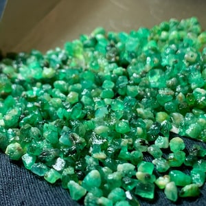 May include: A collection of small, raw emerald gemstones featuring various shades of vibrant green. The stones are irregular in shape and size, displaying a mix of translucent and opaque textures with natural inclusions throughout the pile.