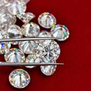 Puede incluir: Colección de diamantes de talla redonda sobre un fondo rojo. Uno de los diamantes es sujetado por unas pinzas, mostrando sus facetas. Los diamantes son transparentes y reflejan la luz, creando un efecto brillante. La imagen resalta el brillo de las gemas.
