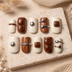 May include: A collection of ten artificial nails with various designs. Some nails are off-white with wooden accents, while others feature silver or gold-colored metal details. The nails are arranged on a neutral-toned surface.