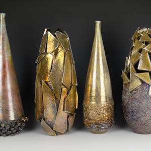 May include: Four decorative vases with distinct designs. The first is copper-toned with a textured base. The second has a gold, fragmented appearance. The third is gold with a textured base. The fourth is textured with gold triangle accents.