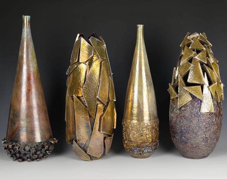 May include: Four decorative vases with distinct designs. The first is copper-toned with a textured base. The second has a gold, fragmented appearance. The third is gold with a textured base. The fourth is textured with gold triangle accents.