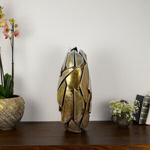 May include: A tall, abstract metal vase with a gold and silver finish. The vase is on a dark wooden surface, next to a potted orchid and a small bouquet of flowers. Books are also visible, creating a decorative display.