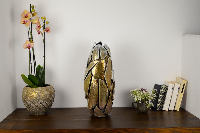 May include: A tall, abstract metal vase with a gold and silver finish. The vase is on a dark wooden surface, next to a potted orchid and a small bouquet of flowers. Books are also visible, creating a decorative display.