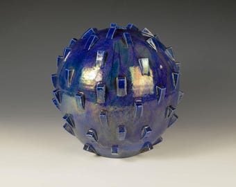 Könnte beinhalten: Eine kugelförmige Keramikskulptur mit tiefblauer, irisierender Glasur. Die Oberfläche ist mit zahlreichen rechteckigen und dreieckigen Vorsprüngen versehen, die ein strukturiertes, stacheliges Aussehen erzeugen. Das Kunstwerk ist ein dekoratives Objekt.