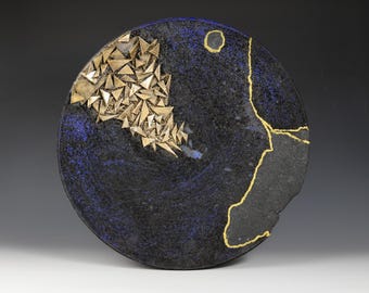 May include: A circular ceramic plate in dark blue with a textured finish. It features a cluster of gold-coloured triangular shapes and a gold-coloured outline. A gold circle is in the upper right corner.