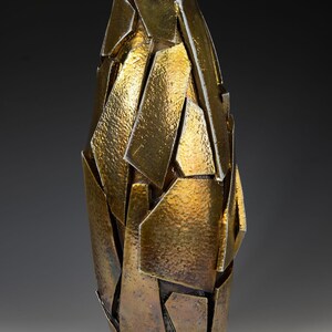 May include: An abstract vase with a metallic gold finish. The vase is made from irregularly shaped, textured pieces, creating a fragmented, geometric design. The overall shape is elongated and slightly tapered, with a height of approximately 30 cm.