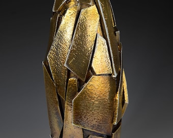 May include: An abstract vase with a metallic gold finish. The vase is made from irregularly shaped, textured pieces, creating a fragmented, geometric design. The overall shape is elongated and slightly tapered, with a height of approximately 30 cm.