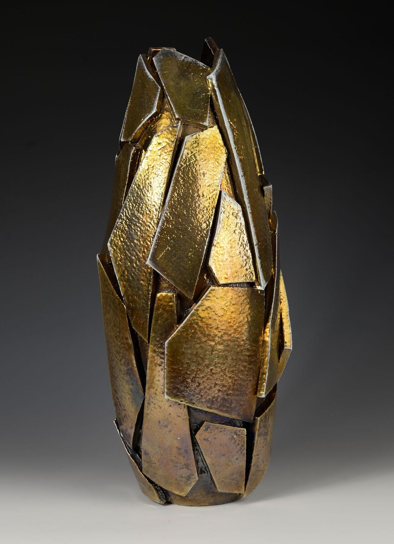 May include: An abstract vase with a metallic gold finish. The vase is made from irregularly shaped, textured pieces, creating a fragmented, geometric design. The overall shape is elongated and slightly tapered, with a height of approximately 30 cm.