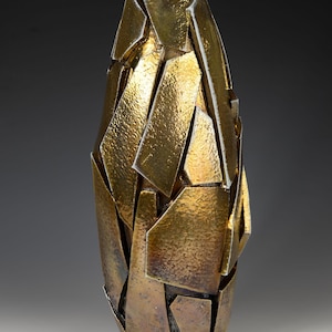 May include: A decorative vase with a unique, fragmented design. The vase is primarily gold with bronze undertones, featuring a textured surface. The abstract shape is reminiscent of shattered pieces, creating a modern aesthetic.