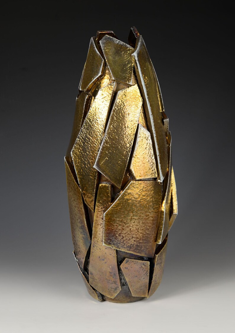 May include: A decorative vase with a unique, fragmented design. The vase is primarily gold with bronze undertones, featuring a textured surface. The abstract shape is reminiscent of shattered pieces, creating a modern aesthetic.
