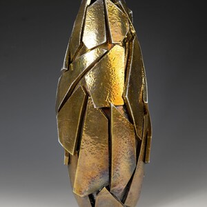 May include: A gold-coloured, abstract vase with a textured surface. The vase is composed of irregularly shaped, overlapping pieces, creating a fragmented appearance. The design gives the impression of a shattered form, with a metallic sheen.