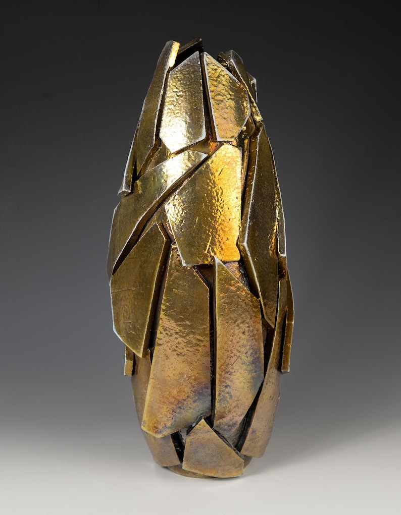 May include: A gold-coloured, abstract vase with a textured surface. The vase is composed of irregularly shaped, overlapping pieces, creating a fragmented appearance. The design gives the impression of a shattered form, with a metallic sheen.