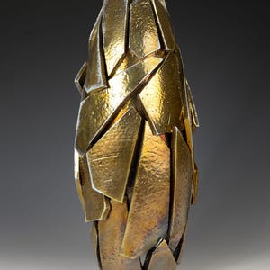 May include: A gold-coloured, abstract sculpture with a fragmented design. The artwork is made up of numerous angular, overlapping shapes, creating a textured surface. The sculpture has a tall, narrow form, and the gold finish gives it a luxurious appearance.