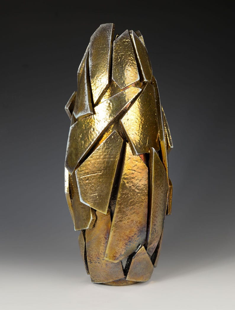 May include: A gold-coloured, abstract sculpture with a fragmented design. The artwork is made up of numerous angular, overlapping shapes, creating a textured surface. The sculpture has a tall, narrow form, and the gold finish gives it a luxurious appearance.