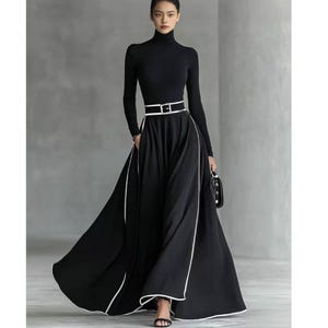 Pode incluir: Um conjunto preto composto por uma blusa de gola alta e uma saia longa preta com detalhes brancos. A saia tem um design amplo e fluido e um cinto branco. O conjunto é complementado com sandálias pretas e uma pequena bolsa preta.