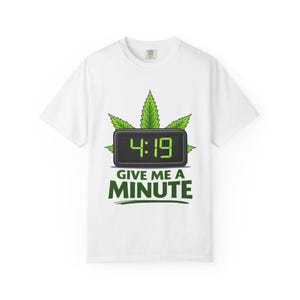 May include: White t-shirt with a green graphic. The graphic shows a digital clock displaying 4:19, framed by green cannabis leaves, and the text "GIVE ME A MINUTE" in green. The shirt has a crew neck.