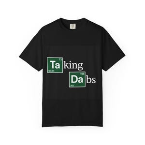 May include: Black t-shirt featuring the words "Taking Dabs" in white. The letters "Ta" and "Da" are in green boxes with chemical element symbols. A simple, short-sleeved tee.