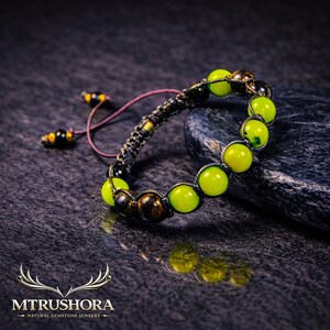 May include: A beaded bracelet featuring alternating bright lime green and dark brown speckled gemstone beads strung on a dark cord. The bracelet includes two adjustable cord ends with small beads and the text MTRUSHORA NATURAL GEMSTONE JEWELRY.