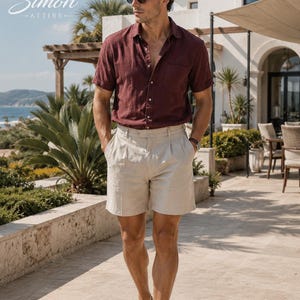 May include: A short-sleeved burgundy button-down shirt with a chest pocket paired with beige pleated shorts and white loafers. The outfit includes black sunglasses and a wristwatch. The text Simon Attire is visible in the upper left corner.