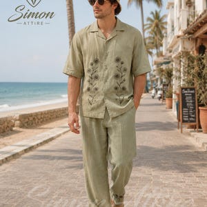 May include: A sage green short-sleeved button-down shirt and matching trousers set featuring black floral embroidery on the shirt front and pant hem. The outfit includes a beige crochet bucket hat and beige slip-on shoes.