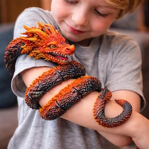 Può includere: Un giocattolo a forma di drago dettagliato, arancione e rosso, si avvolge attorno a un braccio. La testa del drago, con fauci aperte e occhi gialli, è in cima. Il corpo è un mix di arancione, marrone e nero, con squame strutturate. Una persona indossa una maglietta grigia.
