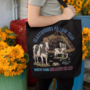May include: Black tote bag with a cow illustration, a barn, and the text "GLASTONBURY FALLOW YEAR" and "WORTHY FARM: RECLAIMING THE LAND". The cows are wearing accessories. The bag is held by a person.