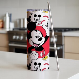 May include: A white tumbler featuring Mickey Mouse in a red outfit, with a stainless steel straw. The tumbler has a repeating pattern of Mickey Mouse, paw prints, and yellow accents. The tumbler is cylindrical.
