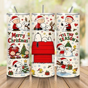 May include: Three cylindrical tumblers with straws featuring a Peanuts holiday theme. The cream-coloured surface displays Snoopy, Charlie Brown, and Woodstock in winter scenes with red doghouses, Christmas trees, snowmen, and the text Merry Christmas and Tis the Season.