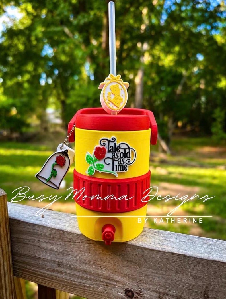 May include: A yellow and red insulated tumbler with a straw and decorative charms. The tumbler features the text "Tale as old as time" and a rose graphic. A charm of a rose in a bell jar is attached.