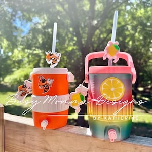 May include: Two drinks dispensers with straws. One is orange with bee accents, the other is a gradient of colours with a lemon slice design. Both have spouts and decorative charms. The text "Busy Momma Designs" and "By Katherine" are visible.
