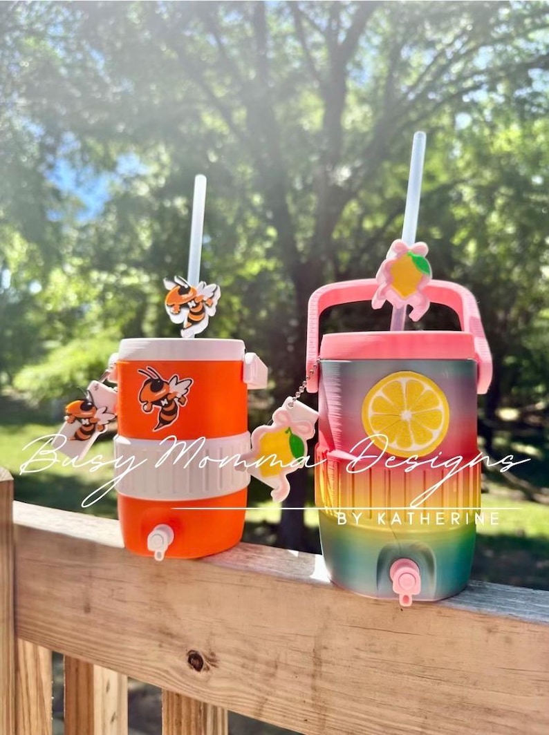 May include: Two drinks dispensers with straws. One is orange with bee accents, the other is a gradient of colours with a lemon slice design. Both have spouts and decorative charms. The text "Busy Momma Designs" and "By Katherine" are visible.