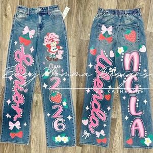 May include: Blue denim jeans with custom painted designs. The front features a Strawberry Shortcake character, pink script, strawberries, bows, and the text "Class of 2026". The back has pink script, strawberries, bows, and the text "NCLA".
