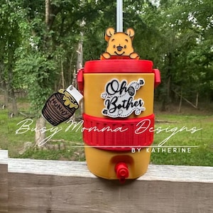 May include: Yellow and red beverage dispenser with a Winnie the Pooh theme. The dispenser has the words "Oh Bother" and a honey pot charm. A Winnie the Pooh figure sits on top. The dispenser has a spigot for dispensing drinks.