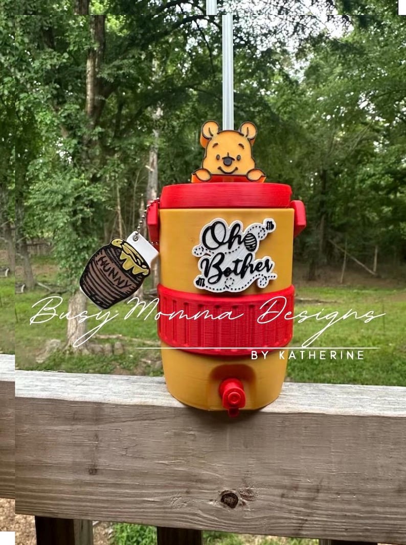 May include: Yellow and red beverage dispenser with a Winnie the Pooh theme. The dispenser has the words "Oh Bother" and a honey pot charm. A Winnie the Pooh figure sits on top. The dispenser has a spigot for dispensing drinks.