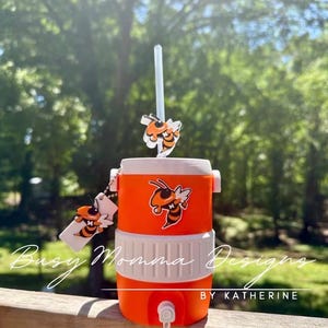 May include: An orange and white cooler with a straw and bee-themed decorations. The cooler has a tap and a bee graphic. The text "Busy Momma Designs" and "BY KATHERINE" are visible on the cooler.