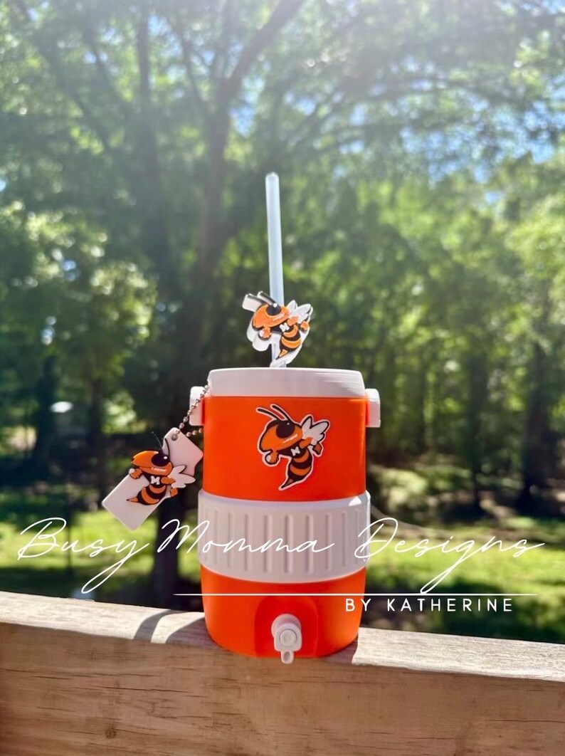 May include: An orange and white cooler with a straw and bee-themed decorations. The cooler has a tap and a bee graphic. The text "Busy Momma Designs" and "BY KATHERINE" are visible on the cooler.