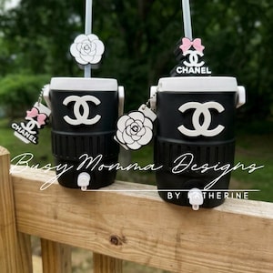 May include: Two black and white insulated tumblers with straws, featuring the Chanel logo and decorative charms. The tumblers have a white top and a black base. The text "Busy Momma Designs by Katherine" is visible.