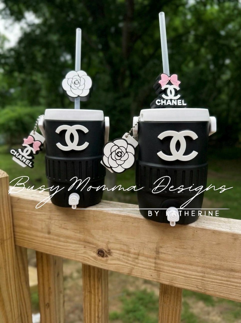 May include: Two black and white insulated tumblers with straws, featuring the Chanel logo and decorative charms. The tumblers have a white top and a black base. The text "Busy Momma Designs by Katherine" is visible.