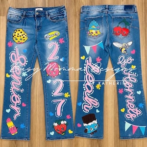 May include: Medium-wash blue denim jeans featuring hand-painted cartoon characters, the text "Senior 2027" on the front, and "Leah" with "Go Hornets" on the back. The design includes colorful hearts, stars, and bunting accents throughout.