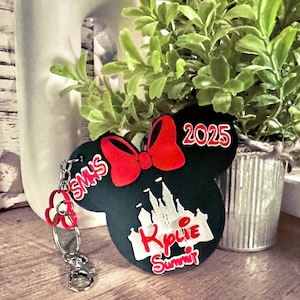 May include: A black Minnie Mouse-shaped keychain with a red bow and the text "2025". The keychain features a castle design and the words "Kylie Summit" in red and white. A red flower charm and silver hardware complete the design.