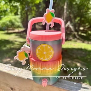 May include: A multi-coloured drinks cooler with a pink lid, handle, and tap. The cooler has a lemon slice graphic and a matching lemon charm. The text "Busy Momma Designs BY KATHERINE" is visible.