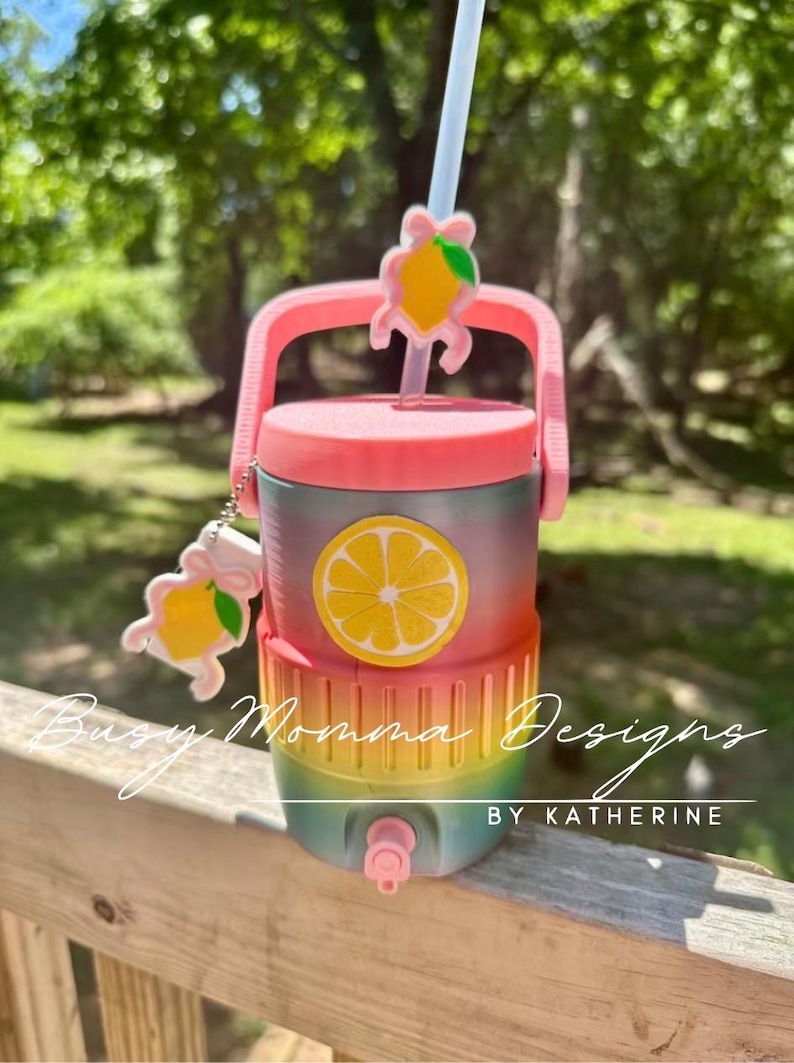 May include: A multi-coloured drinks cooler with a pink lid, handle, and tap. The cooler has a lemon slice graphic and a matching lemon charm. The text "Busy Momma Designs BY KATHERINE" is visible.