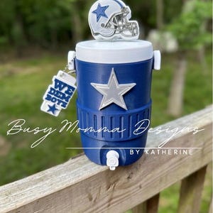 May include: A blue and white water cooler with a silver star and a miniature football helmet on top. A keychain with the text "WE DEM BOYZ" hangs from the side. The cooler is on a wooden surface.