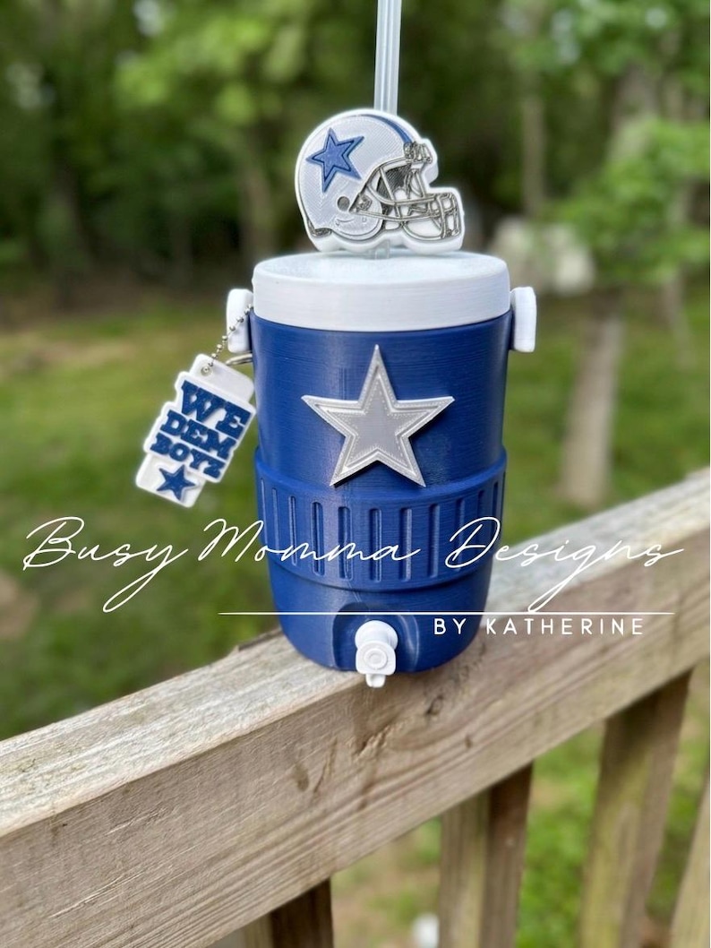 May include: A blue and white water cooler with a silver star and a miniature football helmet on top. A keychain with the text "WE DEM BOYZ" hangs from the side. The cooler is on a wooden surface.