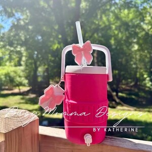 May include: A vibrant pink water cooler with a white handle and a pink bow on the straw. The cooler has a white lid and a tap. The text "Busy Momma Designs by Katherine" is visible on the cooler.