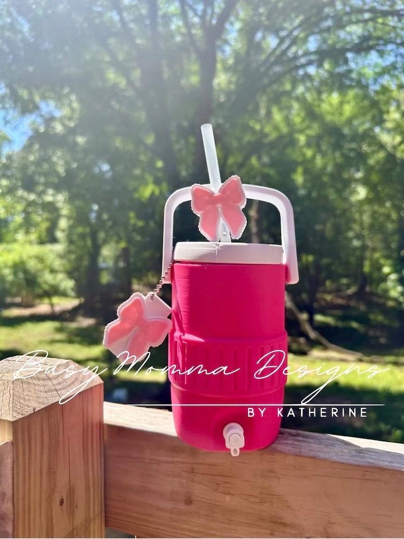 May include: A vibrant pink water cooler with a white handle and a pink bow on the straw. The cooler has a white lid and a tap. The text "Busy Momma Designs by Katherine" is visible on the cooler.