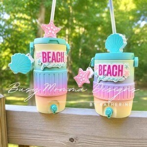 May include: Two beach-themed water coolers with a light blue lid, yellow base, and pastel rainbow centre. Each cooler has a straw, a starfish and seashell charm, and the words "Beach Please". The coolers are ideal for keeping drinks cool.