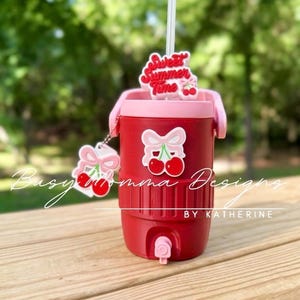 May include: Red insulated cooler with a pink lid and handle, decorated with cherry and bow decals. A straw protrudes from the top, and a cherry charm hangs from the side. The words "Sweet Summer Time" are on a topper.
